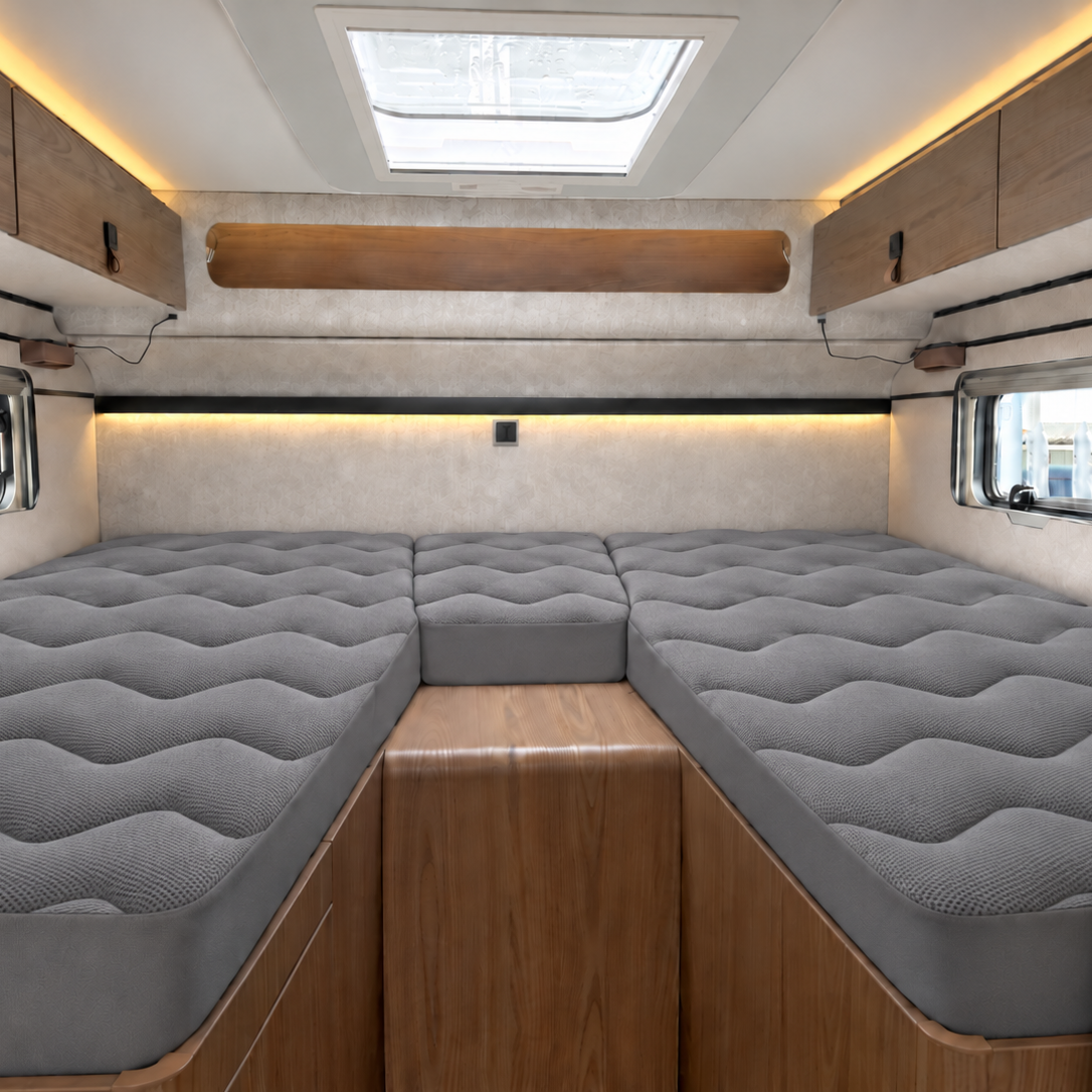 A clean, well-lit interior of a camper van or motorhome showing a U-shaped bed setup with gray quilted mattress toppers, light wood cabinetry, and a skylight.