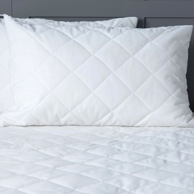 White quilted comforter and pillows on a bed with a nightstand and lamp in the background.