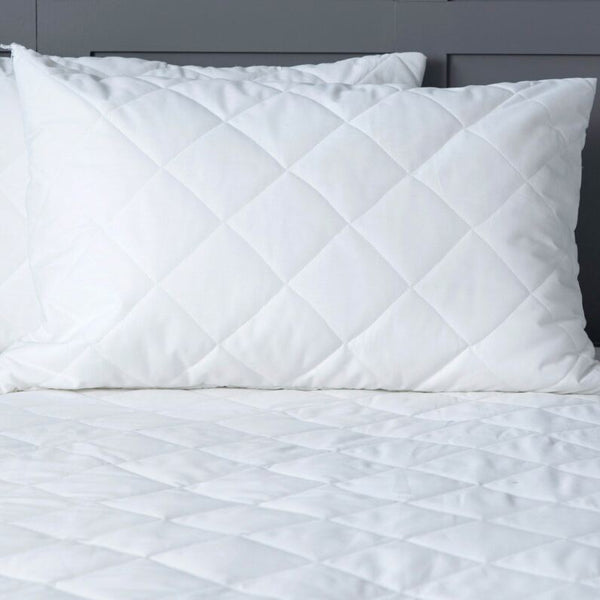 White quilted comforter and pillows on a bed with a nightstand and lamp in the background.